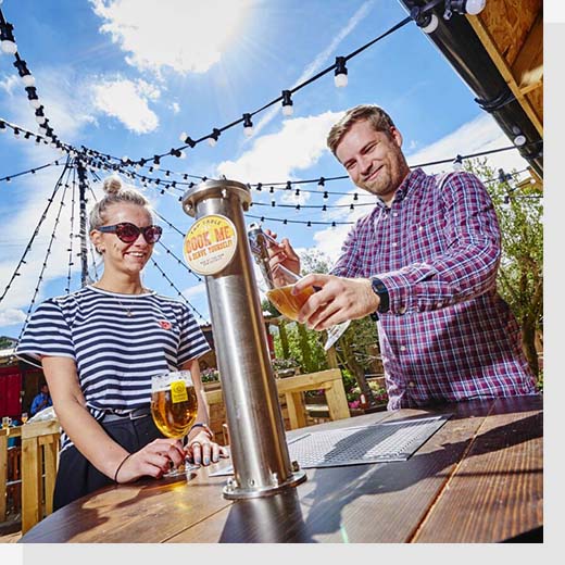 A man and a woman stood in a pub garden on a sunny day, with the man pouring a pint