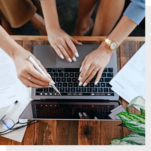 Two pairs of hands, one pointing at a laptop screen and the other using the keyboard on top of a wooden desk 