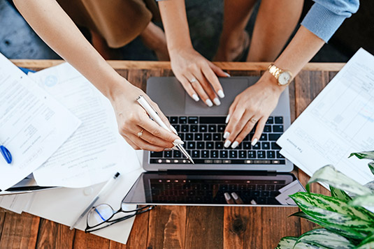 Two pairs of hands using a laptop on top of a wooden desk, one pair of hands pointing at the screen and one pair typing